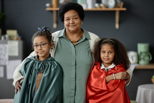 In the image a woman is posing with two young girls both wearing superhero capes.