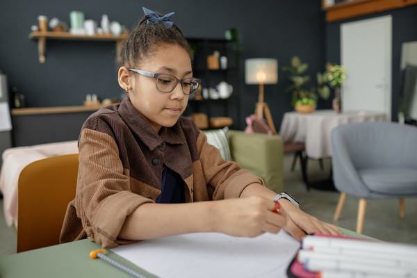 In the image a young girl wearing glasses is sitting at a dining table with a notebook and pencil in front of her.