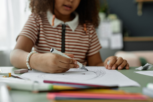 In this image a young girl is sitting at a desk and drawing on a piece of paper with a white pen.