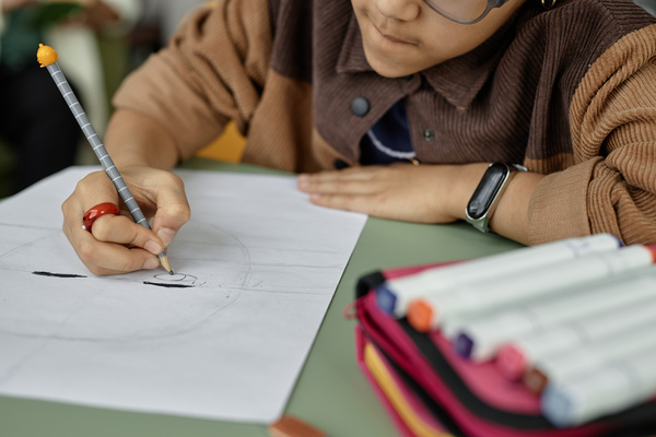 In this image a young boy is sitting at a desk holding a pencil and drawing on a piece of paper.